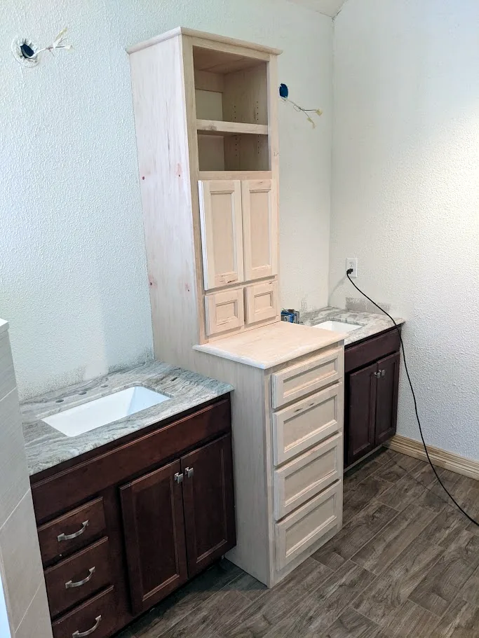 Custom center linen tower and drawer base added between two existing bathroom vanities, shown unpainted before finish, with new wood-look tile floor, Memorial Creek, Spring TX